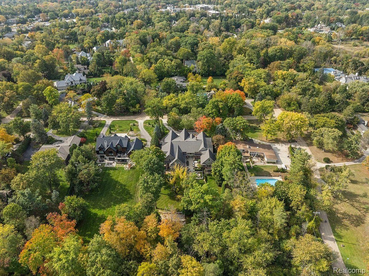 This aerial view showcases a luxurious estate nestled among lush greenery. The property features multiple buildings with dark roofs, manicured lawns, and a swimming pool, suggesting a high-end residential area. The surrounding trees display vibrant fall colors, adding to the property's appeal and creating a sense of privacy and tranquility.