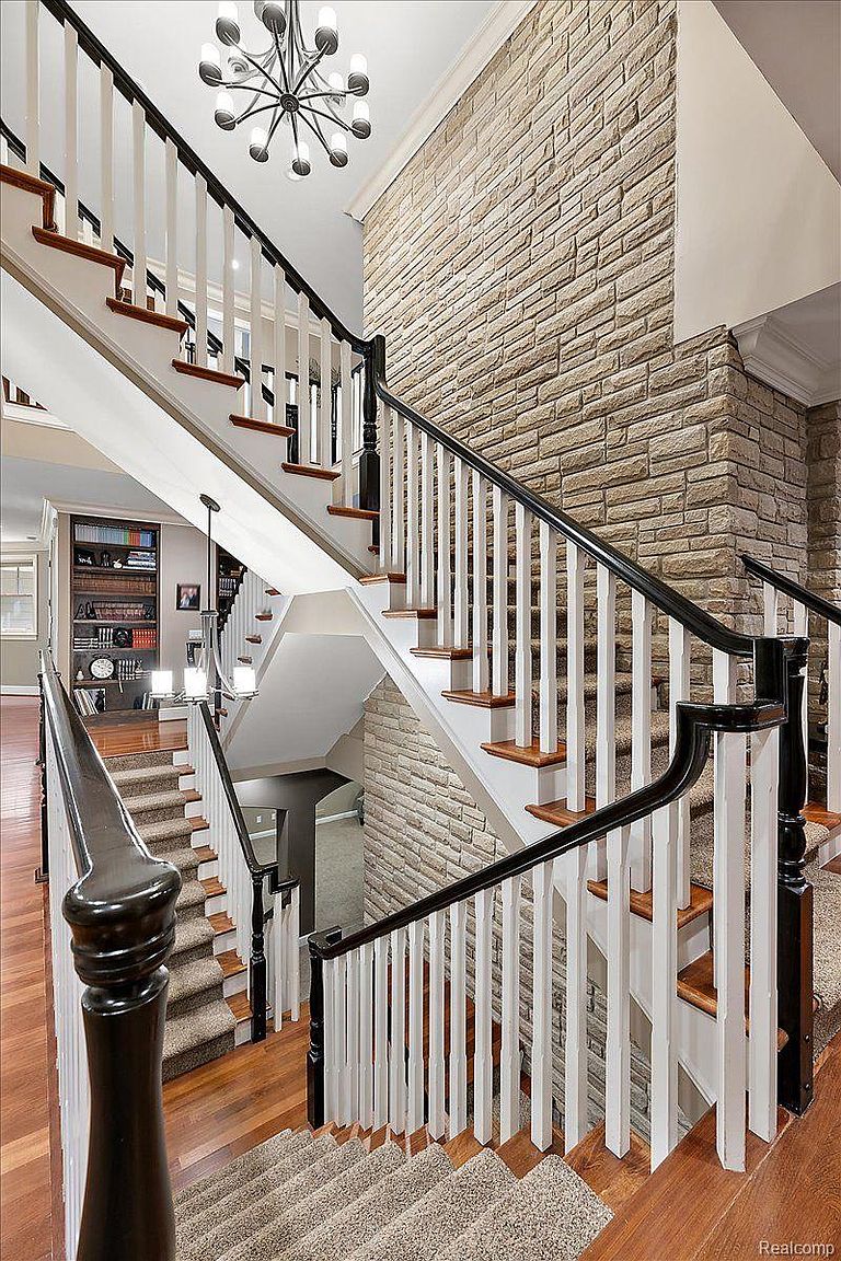 This interior shot showcases a grand staircase with a black and white railing and wooden steps, partially covered with carpet. A textured stone wall adds a sophisticated touch, while a modern chandelier illuminates the space. The staircase leads to different levels, creating a sense of depth and elegance.