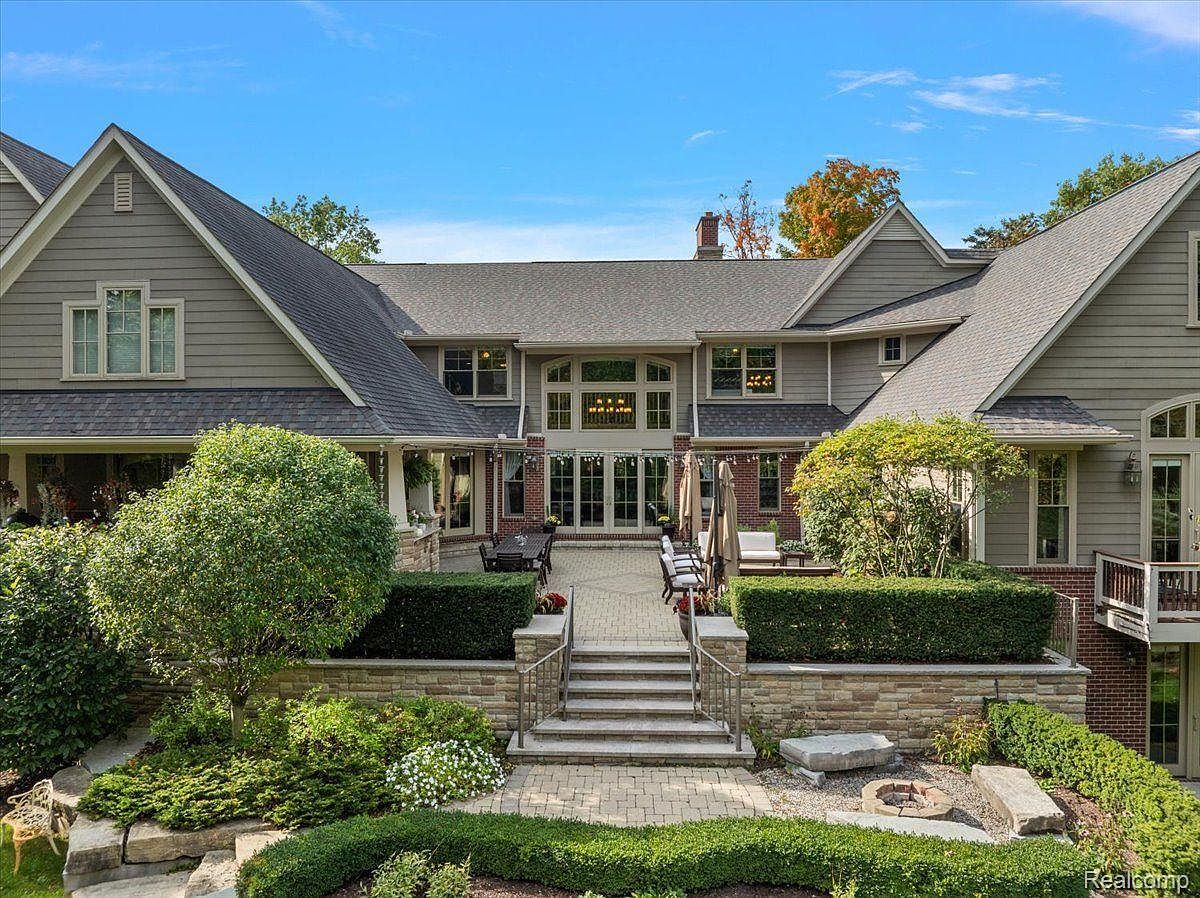 This is a rear view of a large, luxurious home featuring a well-manicured garden and patio area. The house has a complex roofline, multiple windows, and a brick facade on the lower level. The patio is furnished with tables, chairs, and umbrellas, suggesting an inviting outdoor living space.