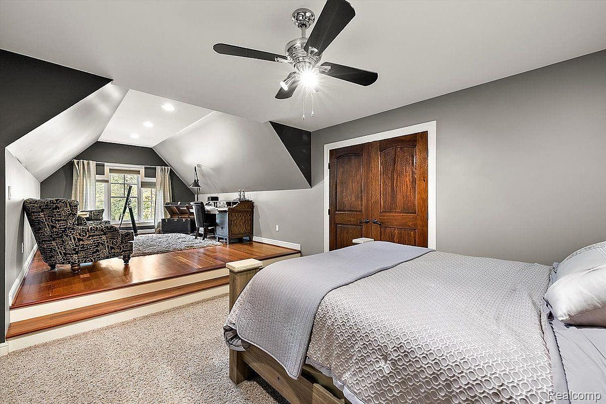 This is a primary bedroom featuring a bed with a gray patterned comforter, a ceiling fan, and a raised platform area with a patterned armchair and a desk. The walls are painted in a neutral gray tone, and the room has a cozy, inviting atmosphere with a mix of textures and materials.