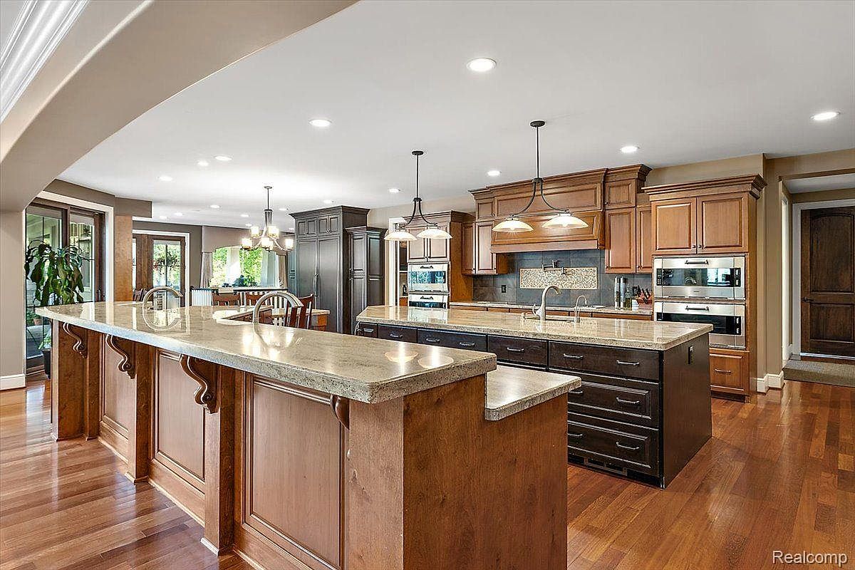 This is a spacious kitchen featuring two islands with light-colored countertops and dark wood cabinetry. Stainless steel appliances are built into the cabinetry, and pendant lights hang above the islands. The hardwood flooring adds warmth to the space, creating an inviting atmosphere.