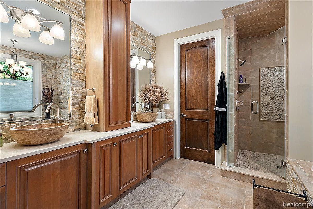 This is a primary bathroom featuring a double vanity with wooden cabinets and vessel sinks. The walls are partially covered with stone tiles, and there's a glass-enclosed shower with tiled walls and a pebble floor. A dark wood door adds a touch of elegance to the space.