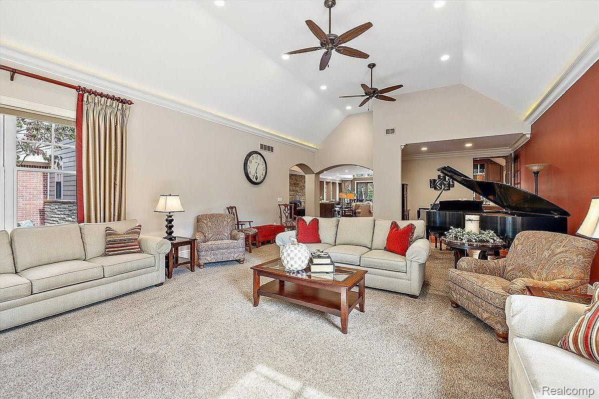 This is an interior shot of a living room featuring two sofas, an armchair, and a grand piano. The room has a high ceiling with two ceiling fans and recessed lighting. The color palette is neutral with pops of red from the throw pillows, creating a warm and inviting atmosphere.