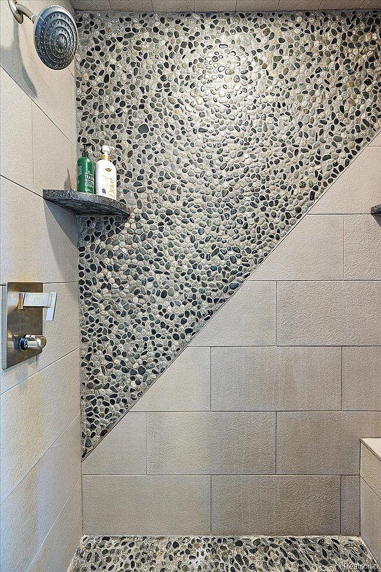 This is a detailed shot of a modern shower featuring a unique design. The shower wall is partially covered with smooth, rounded pebbles, creating a natural and textured accent. The remaining wall space is finished with large, rectangular tiles in a neutral tone, complemented by a built-in shelf and sleek fixtures.