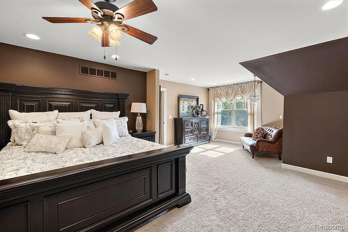 This is a primary bedroom featuring a dark wood, ornate bed frame with numerous pillows, creating a luxurious and inviting sleeping space. The room has a mix of brown and beige tones, with a sitting area near a window providing natural light. The overall impression is one of comfort and elegance.