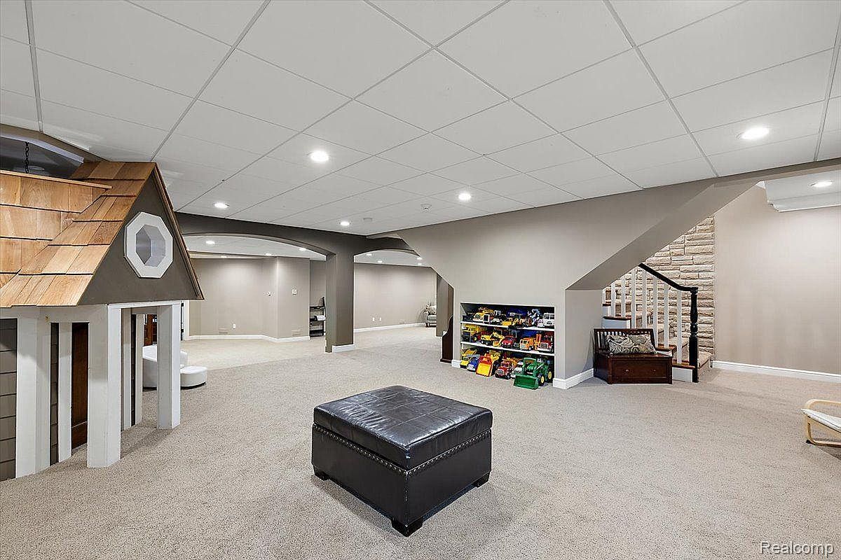 This is a spacious finished basement featuring a play area with a small playhouse, a storage area for toys, and a staircase leading to the upper level. The room is carpeted and has a drop ceiling with recessed lighting. The overall impression is a clean and functional space for recreation and storage.