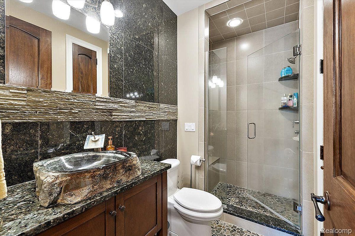 This is a primary bathroom featuring a unique stone vessel sink atop a dark wood vanity with granite countertops. The walls are adorned with dark, textured stone tiles, complemented by a stacked stone accent. A glass-enclosed shower with neutral-toned tiles and pebble flooring adds a modern touch, while the overall design exudes a luxurious and natural aesthetic.