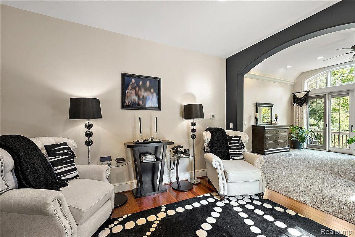 This is an interior shot of a living room featuring two armchairs with black and white pillows, flanking a dark wood side table with lamps. A black and white patterned rug adds a modern touch, while an open archway leads to another area with a dresser and sliding glass doors to the outside. The room is well-lit and appears stylish and comfortable.