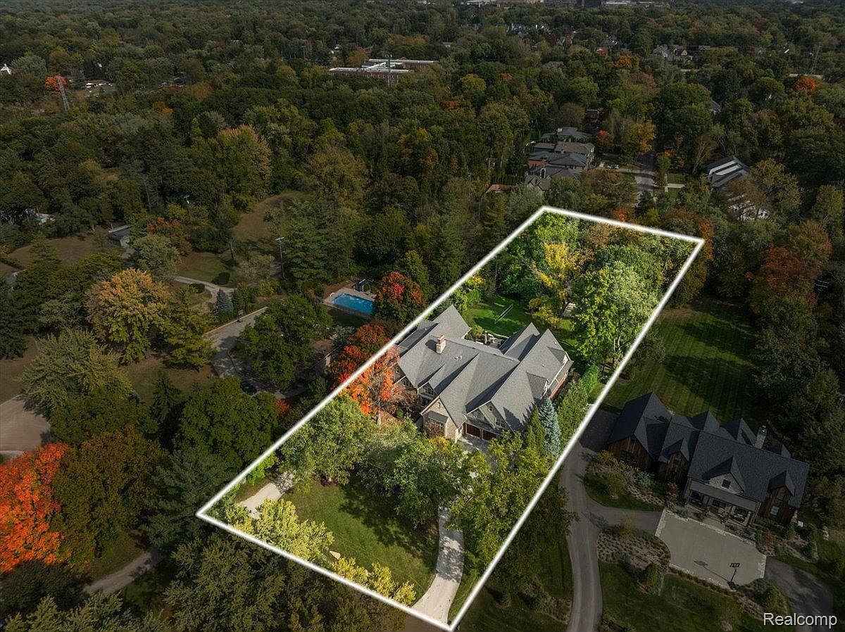 This aerial view showcases a large, luxurious home nestled among mature trees, with a swimming pool visible in the backyard. The property is well-landscaped with a long driveway leading to the house, and a neighboring house is visible to the right. The overall impression is one of privacy and upscale living.