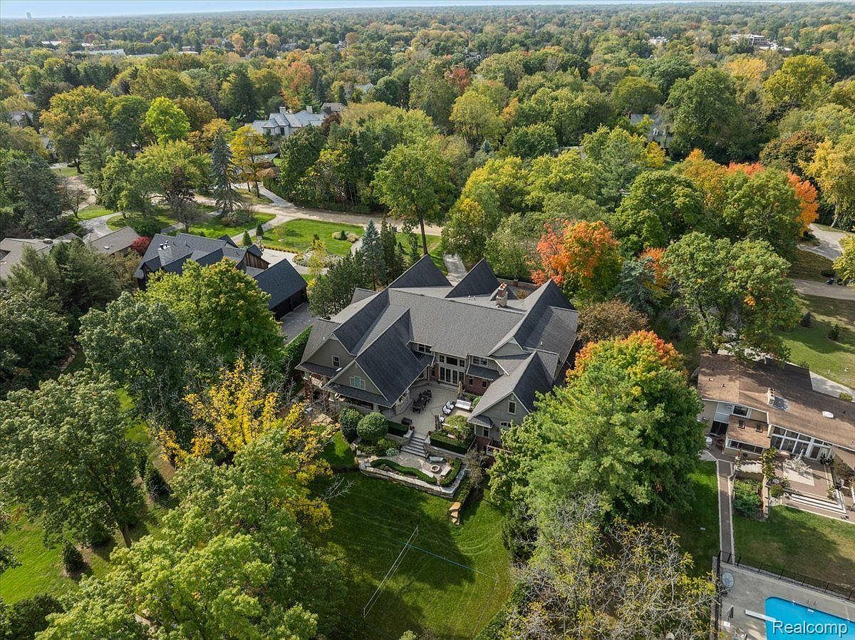 This aerial view showcases a large, luxurious home nestled among mature trees, some displaying fall colors. The house features a complex roofline, a spacious backyard with a patio area, and well-maintained landscaping. The surrounding neighborhood is lush and green, creating a sense of privacy and tranquility.
