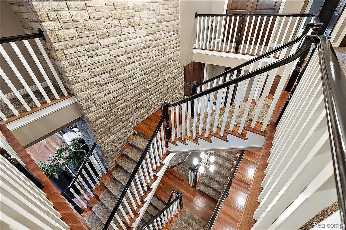 This is an interior shot of a multi-story staircase featuring a combination of hardwood and carpeted steps. The staircase has white balusters and dark wood handrails, creating a classic and elegant look. A stone accent wall adds texture and visual interest to the space, while a chandelier hangs in the lower level, providing ambient lighting.