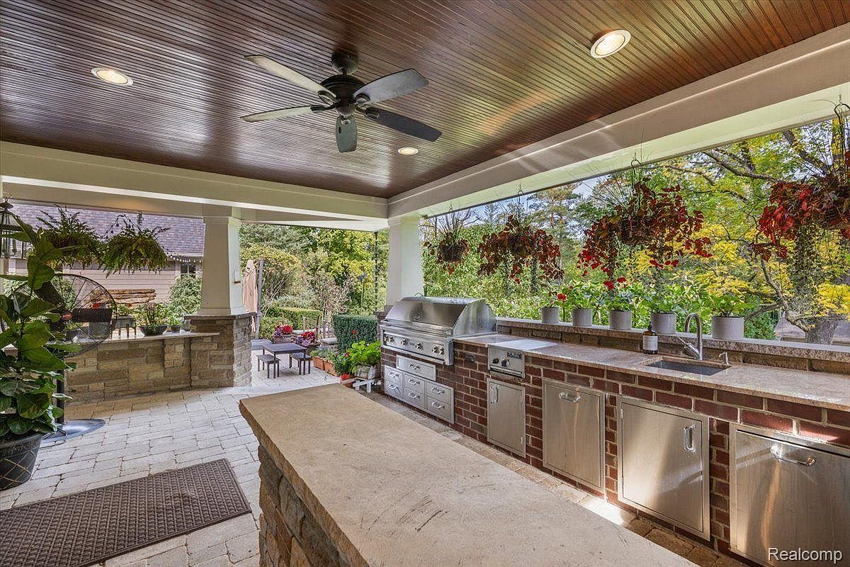 This image showcases a luxurious outdoor patio area, featuring a built-in kitchen with stainless steel appliances and brick accents. The space is covered with a wooden ceiling and a ceiling fan, and the patio is paved with stone. Hanging plants and potted flowers add a touch of nature, while the open design offers views of the surrounding greenery.