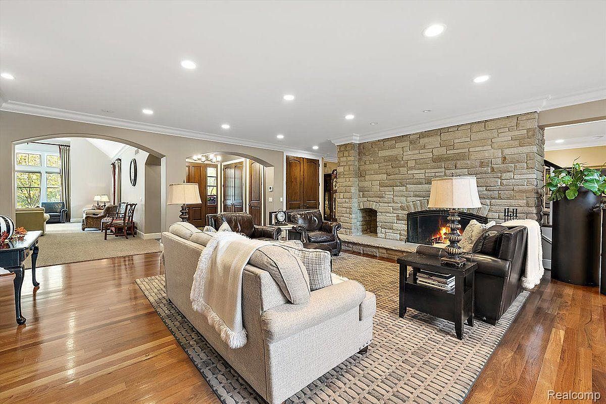 This is an interior shot of a living room featuring a stone fireplace, hardwood floors, and neutral-toned furniture. The room is well-lit with recessed lighting and lamps, creating a warm and inviting atmosphere. The open layout connects to other areas of the home, enhancing the sense of spaciousness.