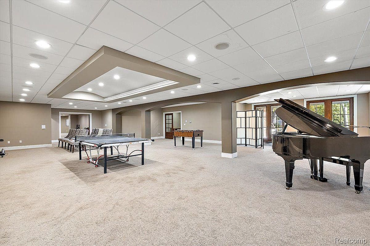 This is a spacious basement featuring a ping pong table, foosball table, and a grand piano. The room is well-lit with recessed lighting and has neutral-colored walls and carpeting. The layout suggests a recreational area, perfect for entertainment and relaxation.