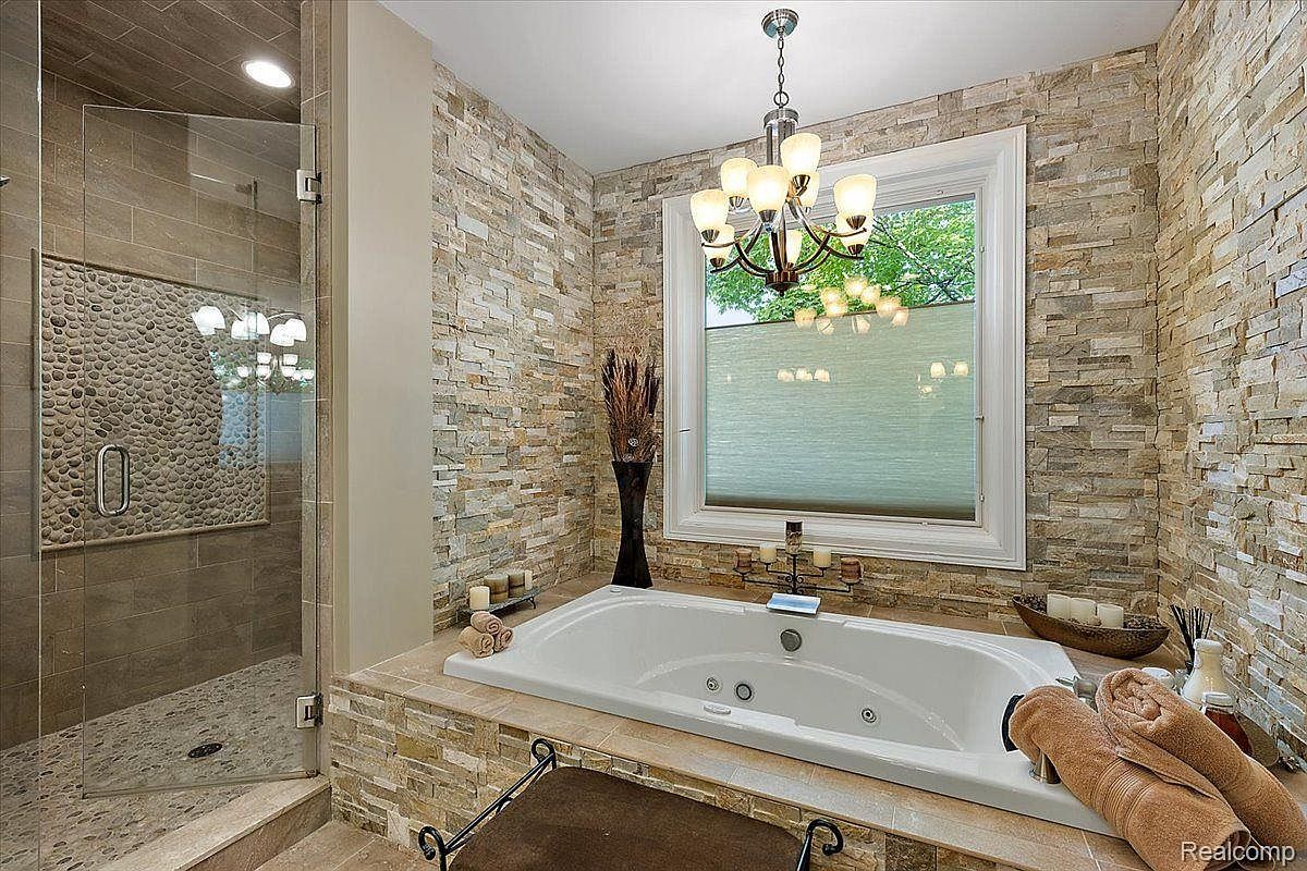 This is a luxurious primary bathroom featuring a large soaking tub surrounded by stone tile, complemented by a modern chandelier. A glass-enclosed shower with pebble accents adds a spa-like feel. The room is well-lit and designed for relaxation, creating a serene and upscale ambiance.