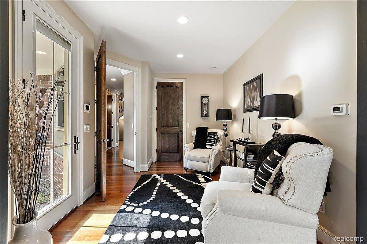 This interior shot showcases a stylish hallway featuring hardwood floors and neutral-toned walls. The space is furnished with two white armchairs, a black and white patterned rug, and side tables with lamps, creating a cozy and inviting atmosphere. A doorway leads to another room, hinting at the home's layout and flow.