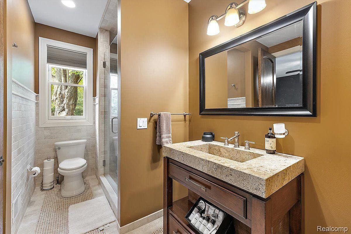 This is a well-lit bathroom featuring a modern vanity with a stone countertop and dark wood cabinetry. A large mirror hangs above the sink, reflecting the room's warm, neutral color scheme. The toilet is positioned near a window, and a glass-enclosed shower is visible, creating a clean and functional space.