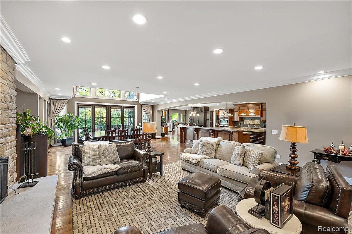 This is an interior shot of a spacious living room featuring two leather sofas, a patterned area rug, and a stone fireplace. The room opens to a dining area and kitchen, creating an open-concept living space. Natural light floods the room through large windows, enhancing the warm and inviting atmosphere.