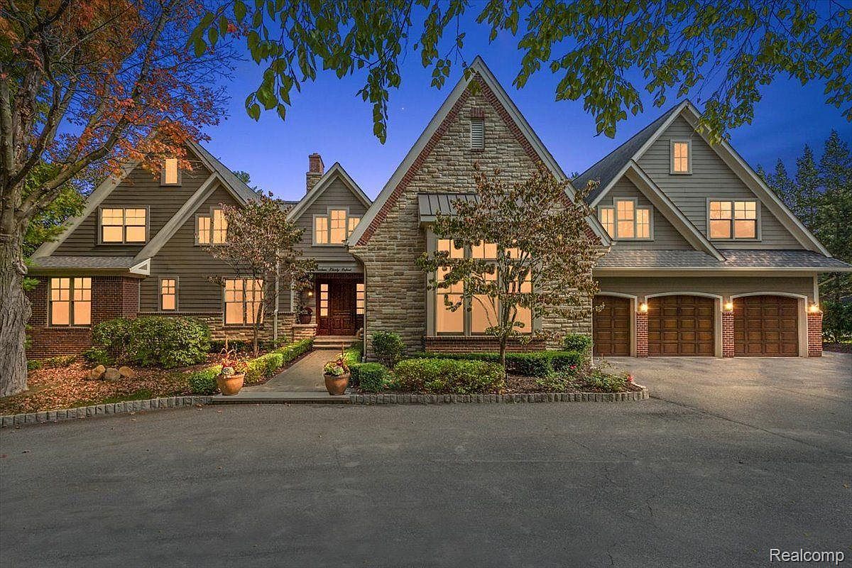 This is a front view of a large, luxurious house with a combination of stone and wood siding. The house features multiple gabled roofs, a three-car garage, and well-manicured landscaping. The driveway is paved and leads to the front entrance, creating an inviting and impressive curb appeal.