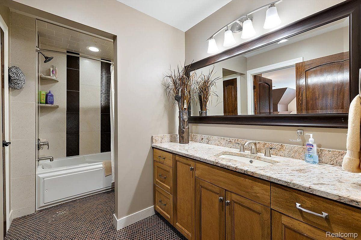 This is a bathroom featuring a vanity with wood cabinets and a granite countertop. A large mirror with a dark frame hangs above the vanity, reflecting a glimpse into another room. The shower/tub area is tiled with a combination of light and dark tiles, and the floor is covered in small, dark mosaic tiles.