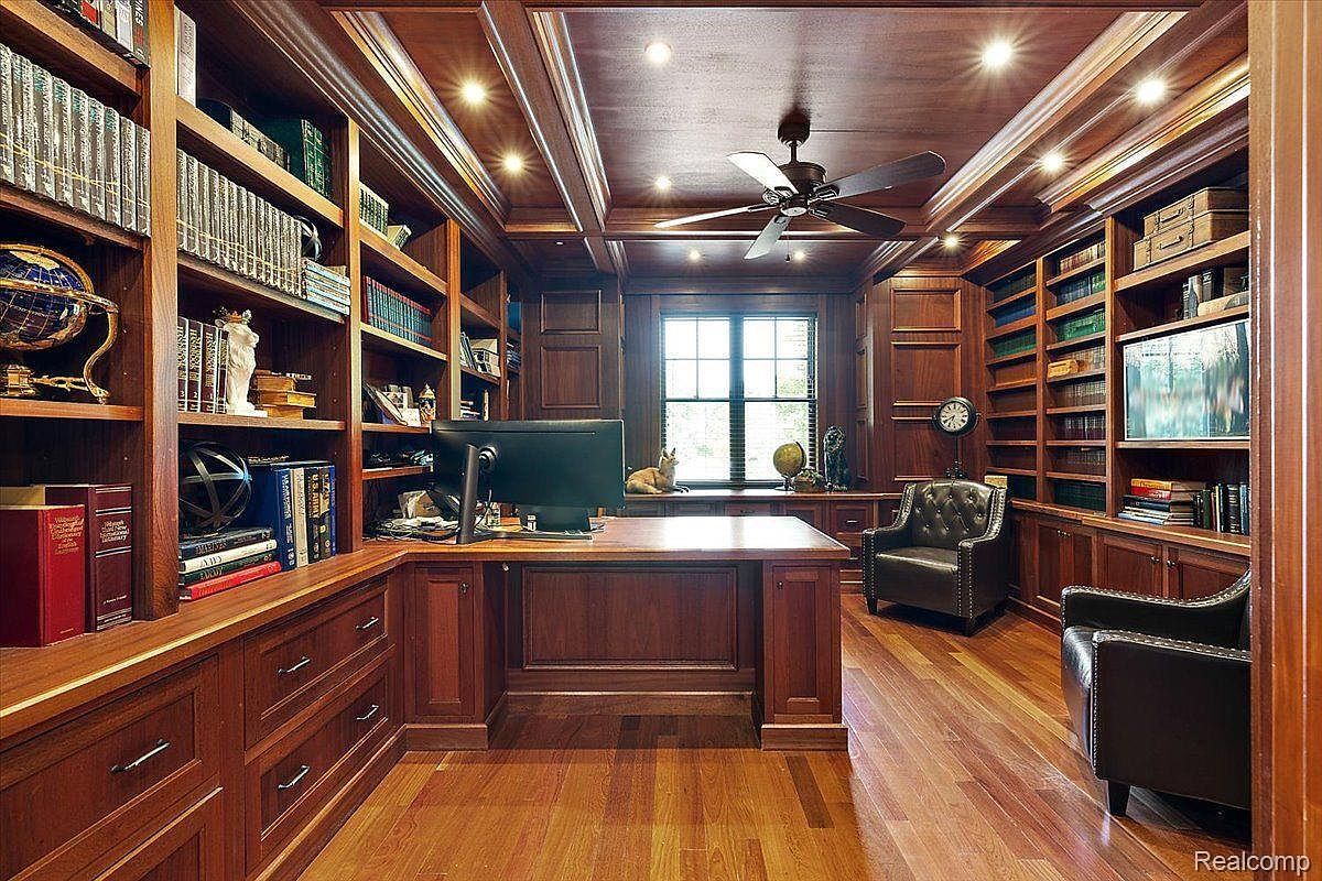 This is a luxurious home office featuring rich wood paneling, built-in bookshelves filled with books and decorative items, and a large wooden desk with a computer. The room is well-lit with recessed lighting and a ceiling fan, creating a warm and inviting atmosphere. Two leather chairs provide comfortable seating, enhancing the room's sophisticated and professional appeal.