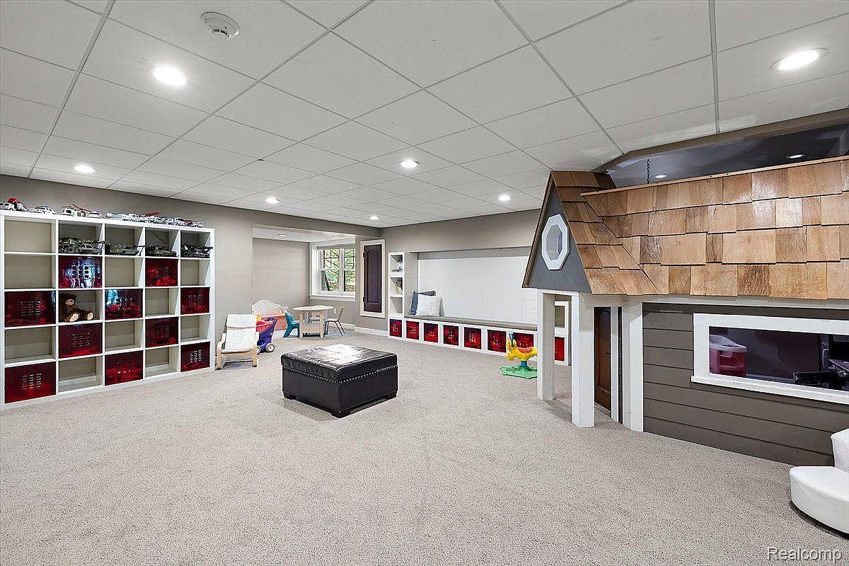 This is a spacious basement area designed as a playroom. It features a large white shelving unit with red storage bins, a black leather ottoman, a playhouse structure with a shingled roof, and a built-in window seat with storage. The room is well-lit with recessed lighting and has neutral-colored carpeting, creating a comfortable and inviting space for children.