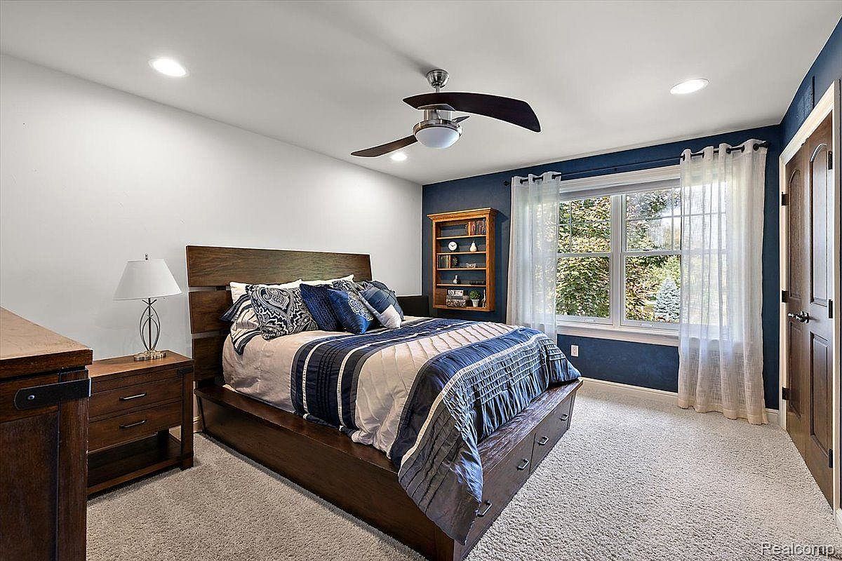 This is a primary bedroom featuring a large bed with a wooden frame and a blue and white patterned comforter. The room has a combination of white and blue walls, a ceiling fan, and a window with sheer curtains. A wooden bookshelf and a nightstand with a lamp are also visible, creating a cozy and inviting atmosphere.