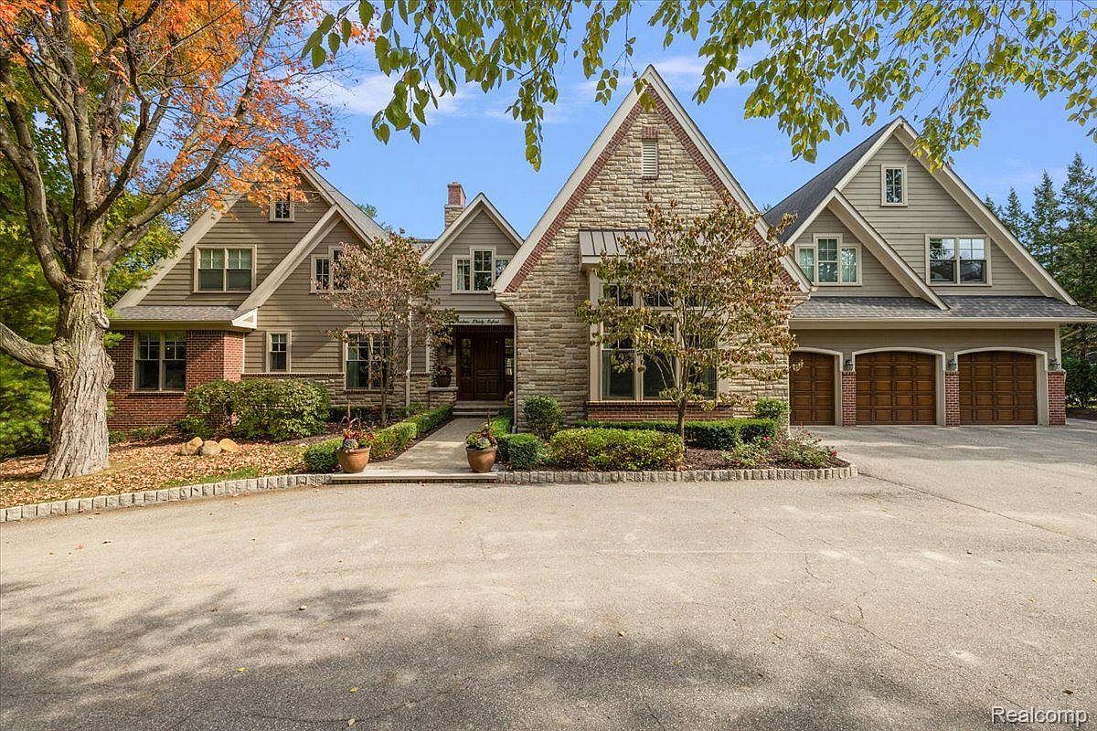 This is a front exterior view of a large, luxurious home. The house features a combination of stone and siding, with multiple gabled roofs and a three-car garage. The landscaping is well-maintained, with mature trees and manicured shrubs, creating a welcoming and upscale impression.