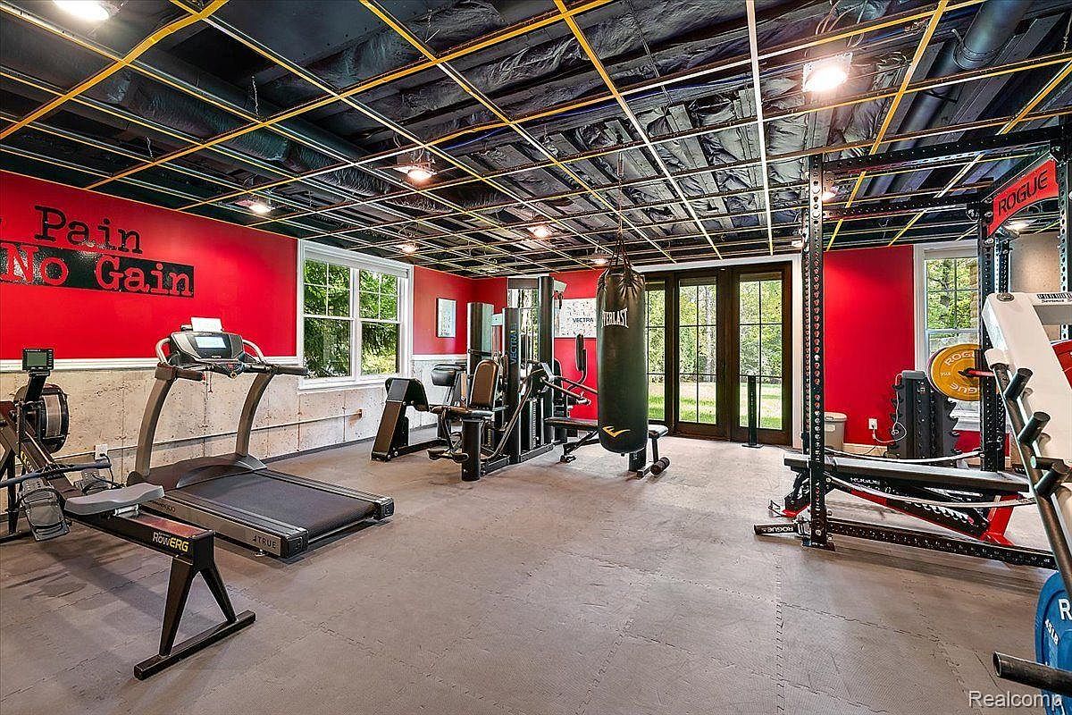 This is a well-equipped home gym featuring a variety of exercise machines including a treadmill, rowing machine, weightlifting rack, and a punching bag. The room has a red accent wall with the phrase "Pain No Gain" and a black ceiling with exposed ductwork. The flooring is a gray rubber mat, providing a safe and comfortable workout surface.