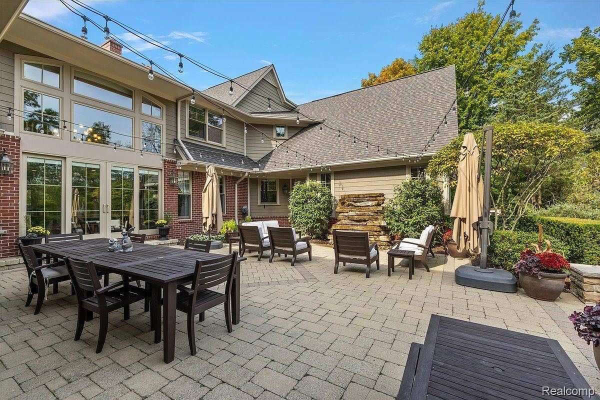 This is a well-appointed outdoor patio area, perfect for entertaining. It features a large dining table with seating for eight, several lounge chairs arranged for conversation, and a large umbrella for shade. The patio is paved with brick pavers and is surrounded by lush landscaping, creating a private and inviting outdoor space.