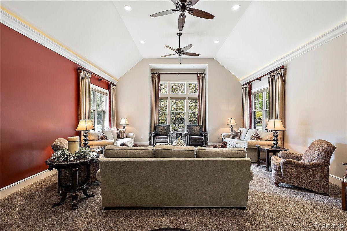 This is an interior shot of a living room featuring a neutral color palette with a pop of red on one wall. The room is furnished with sofas, armchairs, and decorative tables, and the windows are dressed with elegant curtains. Two ceiling fans are present, and the room is well-lit with natural light and lamps, creating a warm and inviting atmosphere.