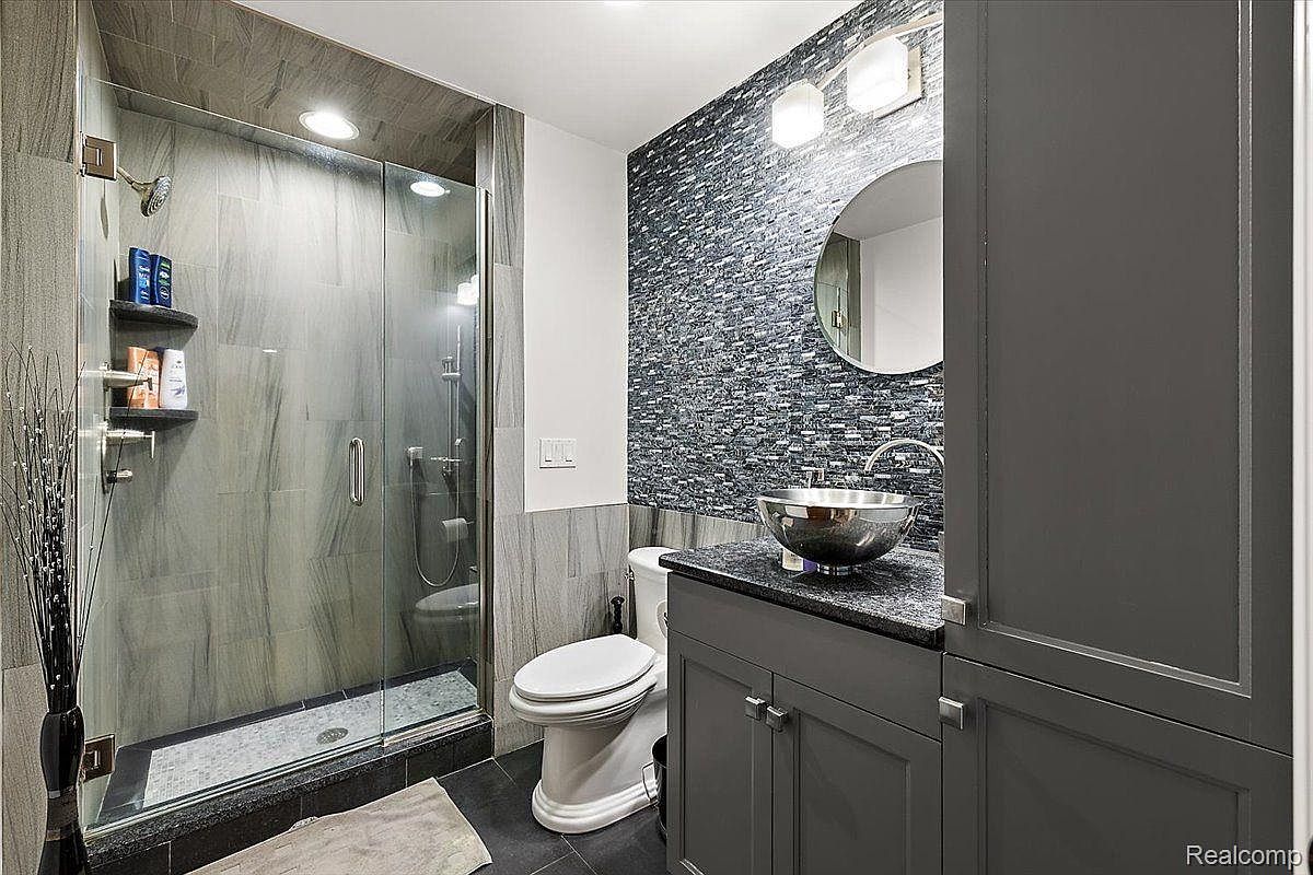 This is a modern bathroom featuring a glass-enclosed shower with gray marble-like tiling, a unique vessel sink on a dark countertop, and a mosaic tile accent wall. The bathroom also includes a toilet and gray cabinetry, creating a sleek and contemporary design. The overall impression is clean and stylish.