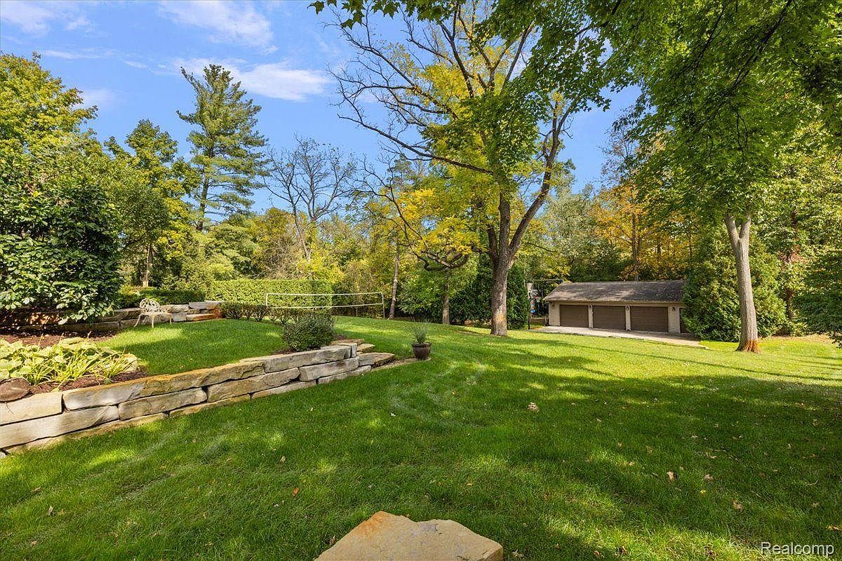 This image showcases a well-maintained backyard with lush green grass, mature trees, and a stone retaining wall. A volleyball net is set up in the distance, and a three-car garage is visible beyond the trees. The yard offers a serene and private outdoor space, perfect for relaxation and recreation.