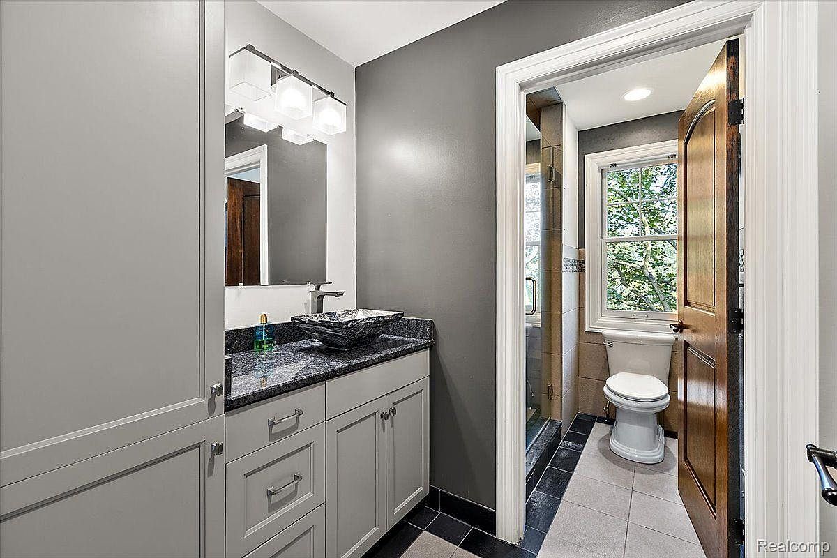This is a bathroom featuring a gray color scheme with a modern vanity, a unique vessel sink, and a granite countertop. The room includes a toilet visible through an open doorway, suggesting an en-suite layout with a shower area. The overall impression is clean and stylish, with a focus on contemporary design elements.