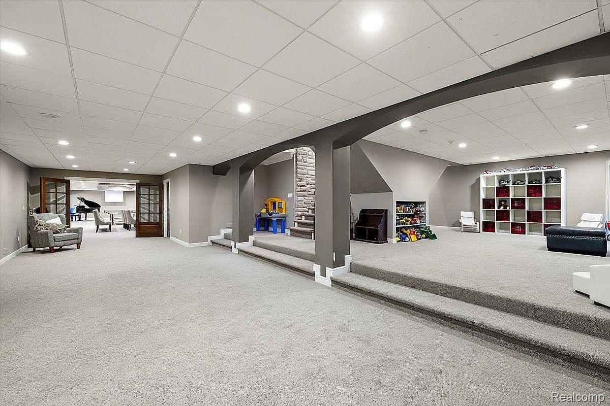 This is a spacious finished basement featuring a large open area with gray carpeting and walls. The room is divided into zones by slightly elevated platforms, creating distinct spaces for different activities. Recessed lighting provides ample illumination, and there are various furniture pieces and storage solutions, suggesting a versatile and family-friendly environment.