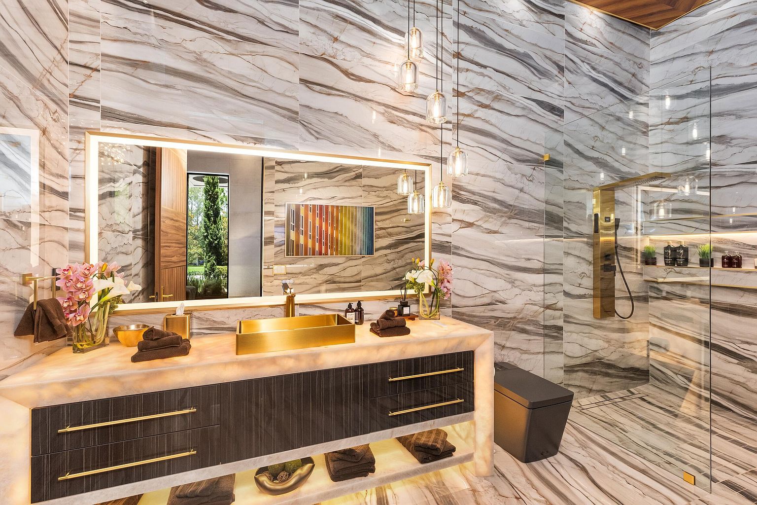This luxurious primary bathroom features striking, large-format patterned stone walls and flooring that create a cohesive, high-end aesthetic. The vanity is highlighted by a glowing, translucent stone countertop, a metallic rectangular sink, and dark cabinetry with gold-toned hardware, all illuminated by a large, backlit mirror and elegant pendant lights. A glass-enclosed shower area with gold fixtures and a dark, modern toilet complete the sophisticated, spa-like design.