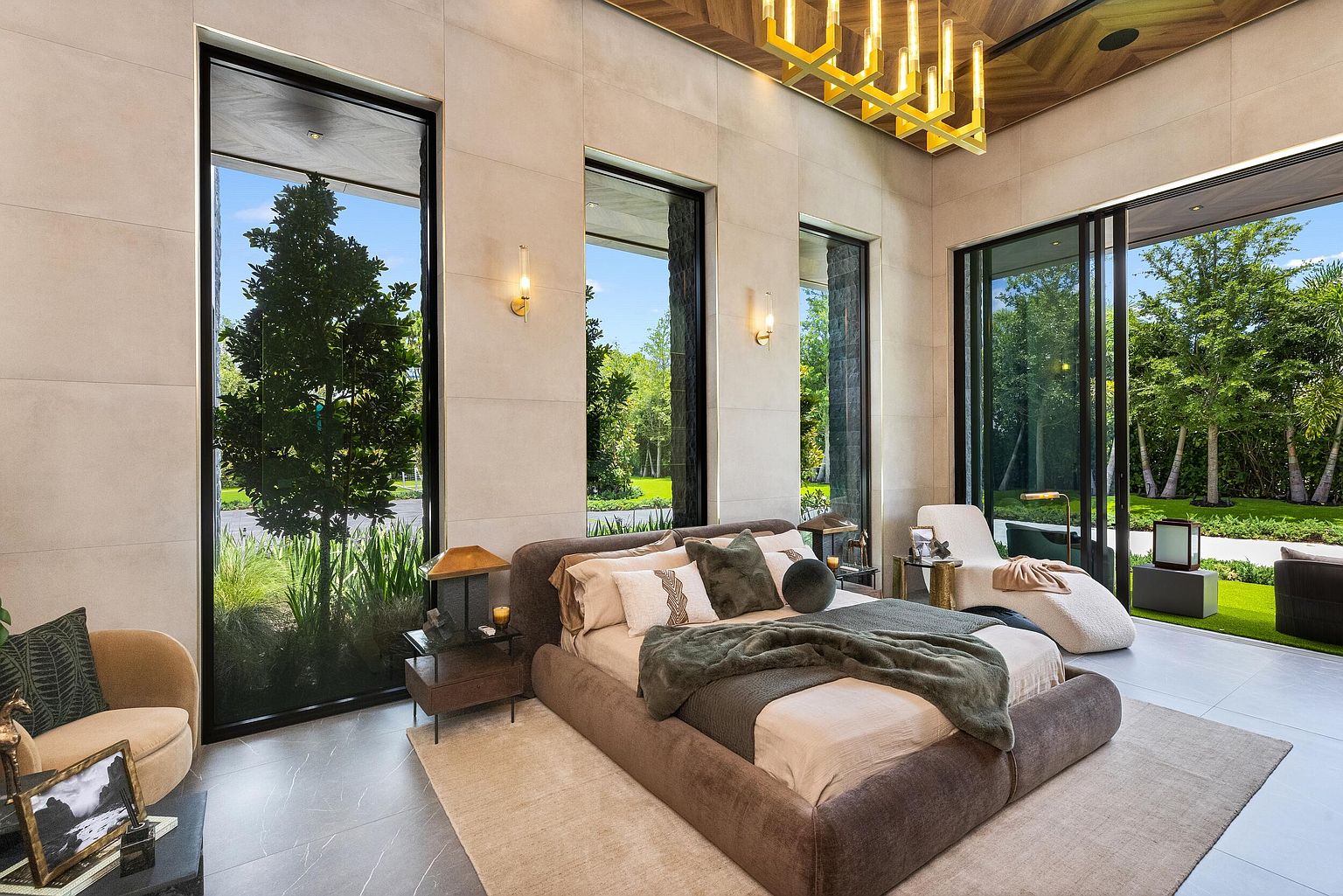 This luxurious primary bedroom features a contemporary aesthetic with a low-profile upholstered bed as the central focus. The room is defined by expansive floor-to-ceiling windows that provide abundant natural light and seamless views of the lush outdoor greenery. A sophisticated gold-toned chandelier hangs from a warm wood-paneled ceiling, complementing the neutral stone-tiled walls and the serene, high-end atmosphere.