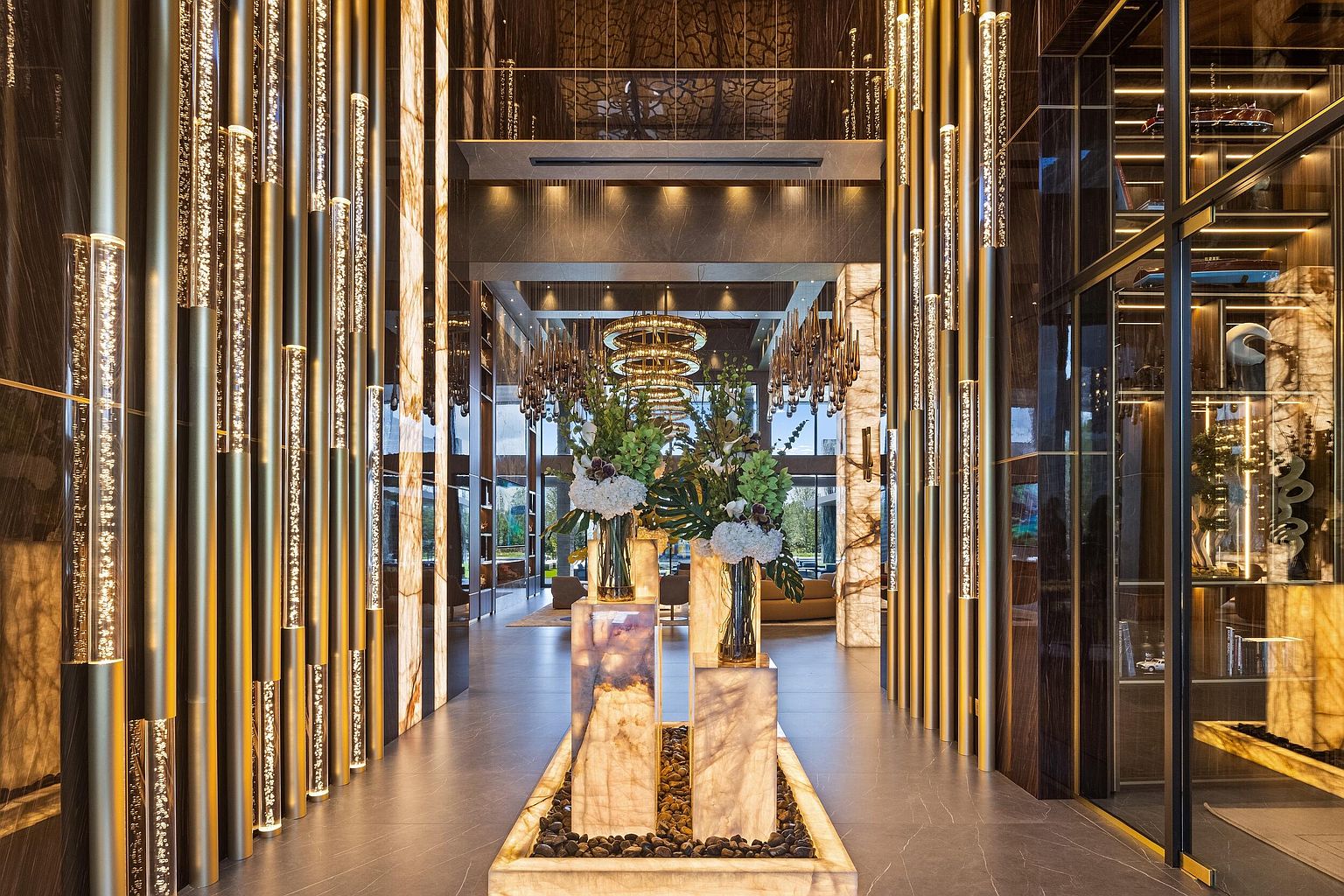 This grand, opulent entryway features a dramatic central display of floral arrangements set atop illuminated stone pedestals surrounded by a bed of dark river stones. The space is defined by a series of vertical, glowing light columns and dark, reflective wall panels that create a sophisticated, high-end atmosphere. The perspective is centered and symmetrical, drawing the eye through the hallway toward a distant, brightly lit living area with a large, intricate chandelier.