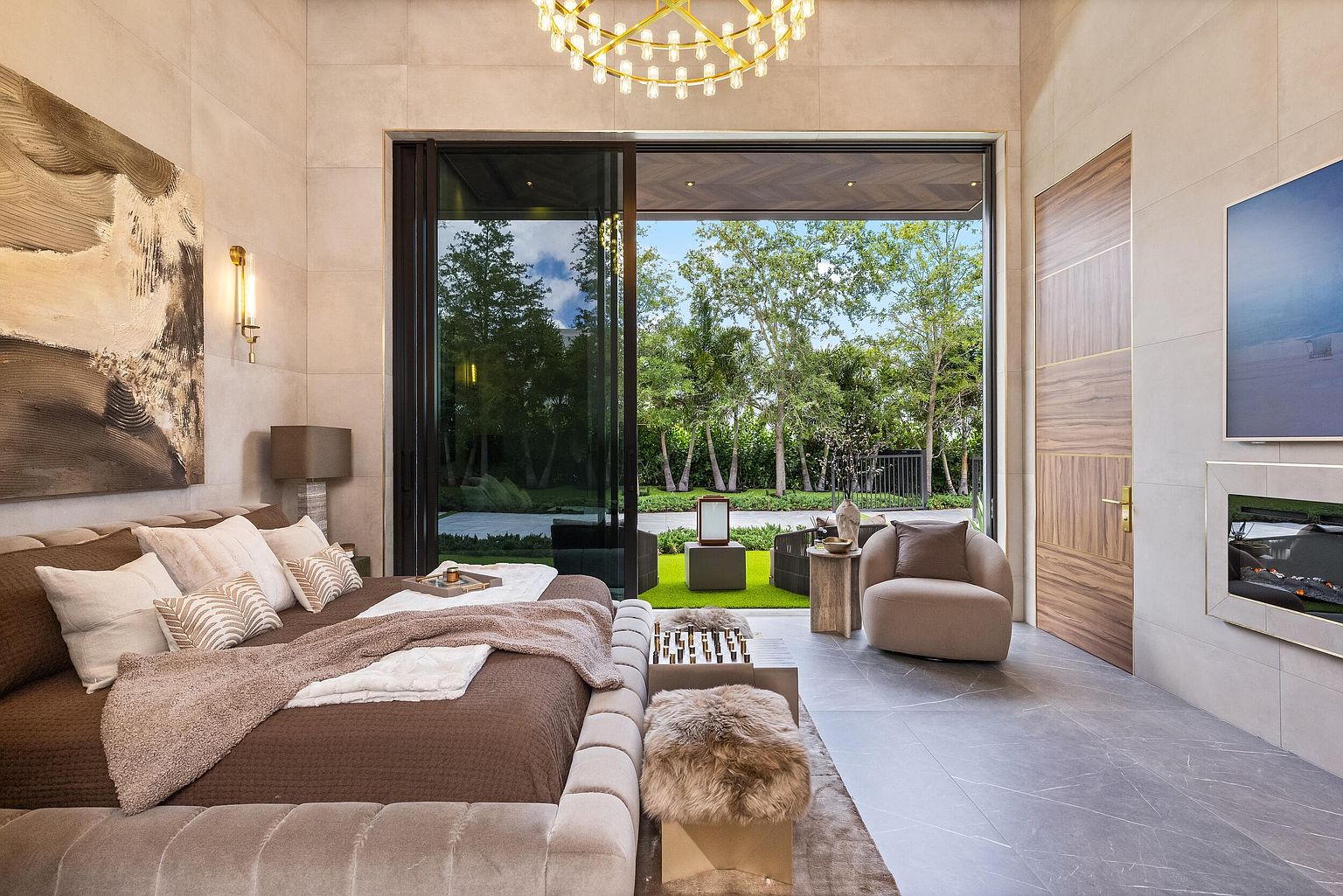 This luxurious primary bedroom features a plush, upholstered bed frame and a neutral color palette that emphasizes comfort and sophistication. Large sliding glass doors open to a lush, green outdoor space, seamlessly blending indoor and outdoor living. The room is accented by a modern chandelier, a cozy armchair, and a sleek fireplace integrated into the wall, creating an inviting and high-end atmosphere.
