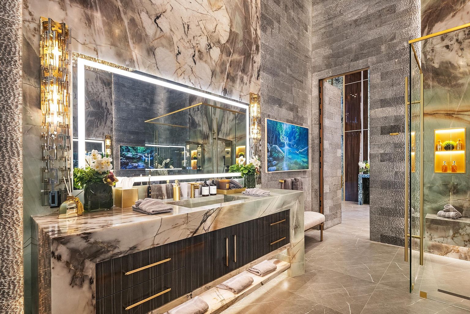 This luxurious primary bathroom features a striking vanity with a thick, patterned stone countertop and dark, textured cabinetry with gold-toned hardware. A large, illuminated mirror serves as the focal point, flanked by ornate, vertical light fixtures, while a glass-enclosed shower area with integrated shelving is visible to the right. The space is finished with large-format stone wall cladding and flooring, creating a sophisticated and high-end atmosphere.