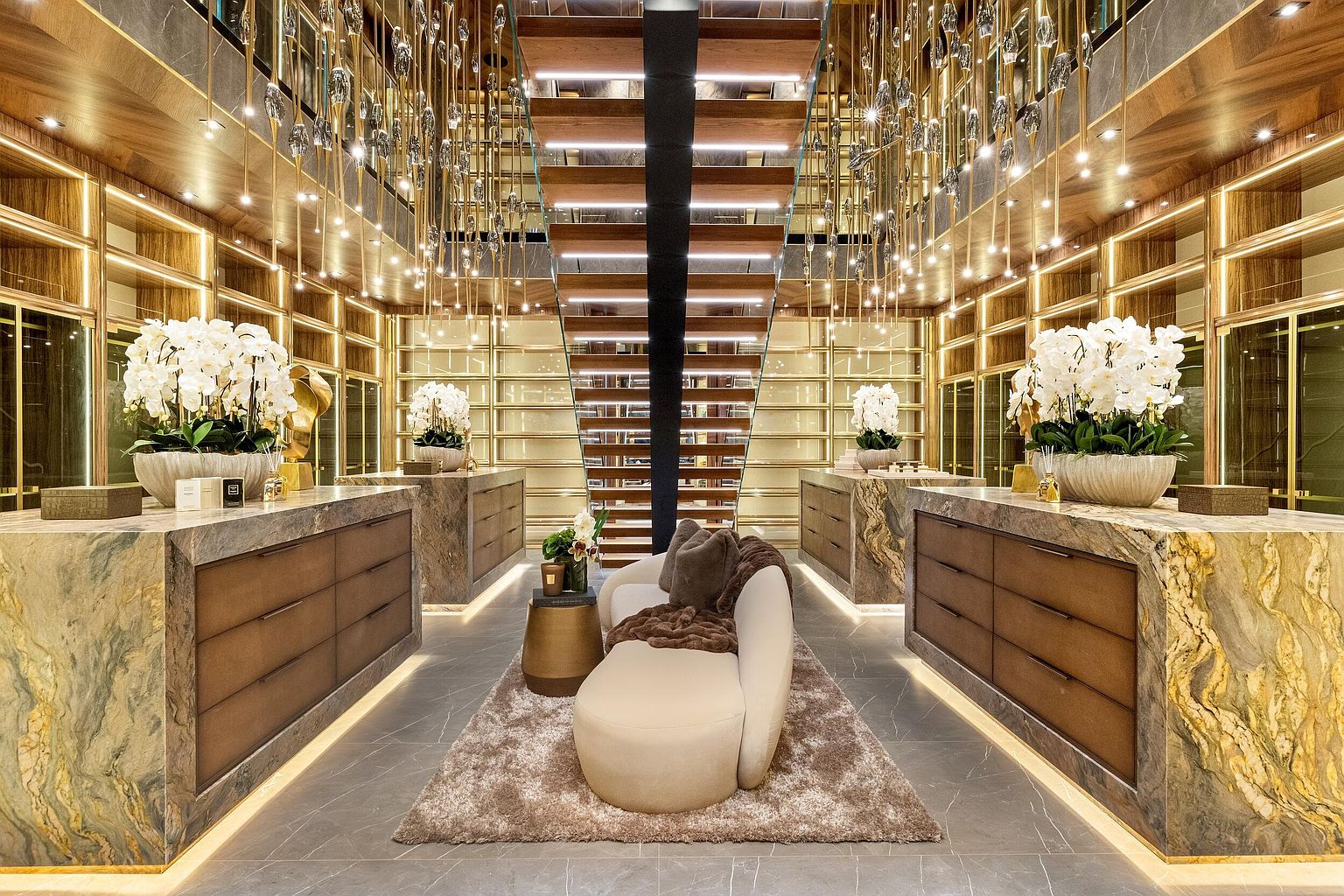 This luxurious walk-in closet features a symmetrical layout with two large stone-topped islands, each adorned with white floral arrangements. A central curved sofa sits on a plush rug, positioned directly in front of a striking staircase with illuminated wooden treads. The space is framed by extensive shelving and cabinetry, all accented by dramatic, cascading pendant lighting that creates a sophisticated and opulent atmosphere.