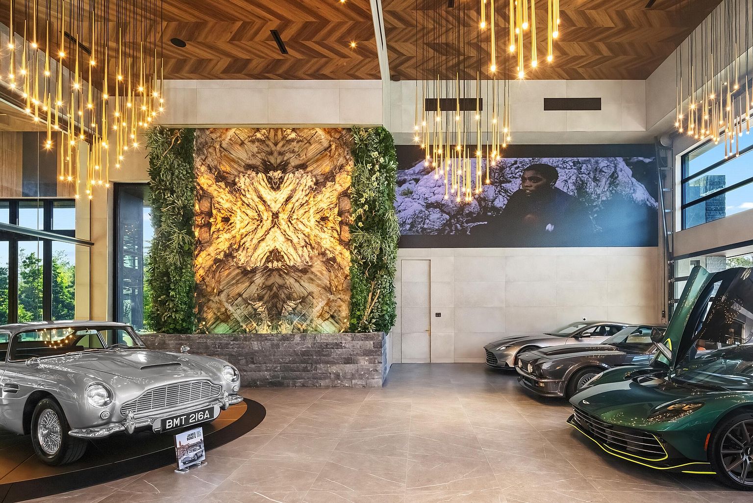 This expansive, high-end garage serves as a sophisticated showroom for a collection of luxury vehicles, including a classic silver sports car and a modern green supercar. The space features a striking illuminated stone feature wall flanked by vertical greenery, a herringbone-patterned wood ceiling, and dramatic cascading pendant lighting. A large mural of a person on the wall adds a cinematic, gallery-like atmosphere to this meticulously designed automotive display area.
