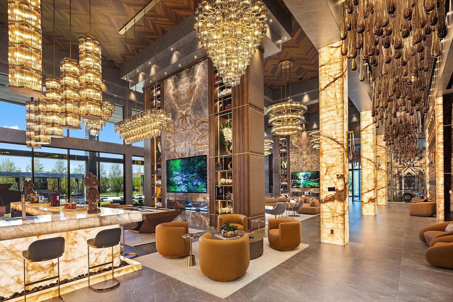 This expansive, high-end lounge area features a sophisticated open-concept design with a backlit stone bar and multiple seating arrangements. The space is defined by dramatic, tiered crystal chandeliers, a tall stone-clad chimney breast housing a television, and large windows that provide a seamless connection to the outdoors. The combination of warm ambient lighting, rich wood accents, and polished stone surfaces creates an atmosphere of refined luxury and grand scale.