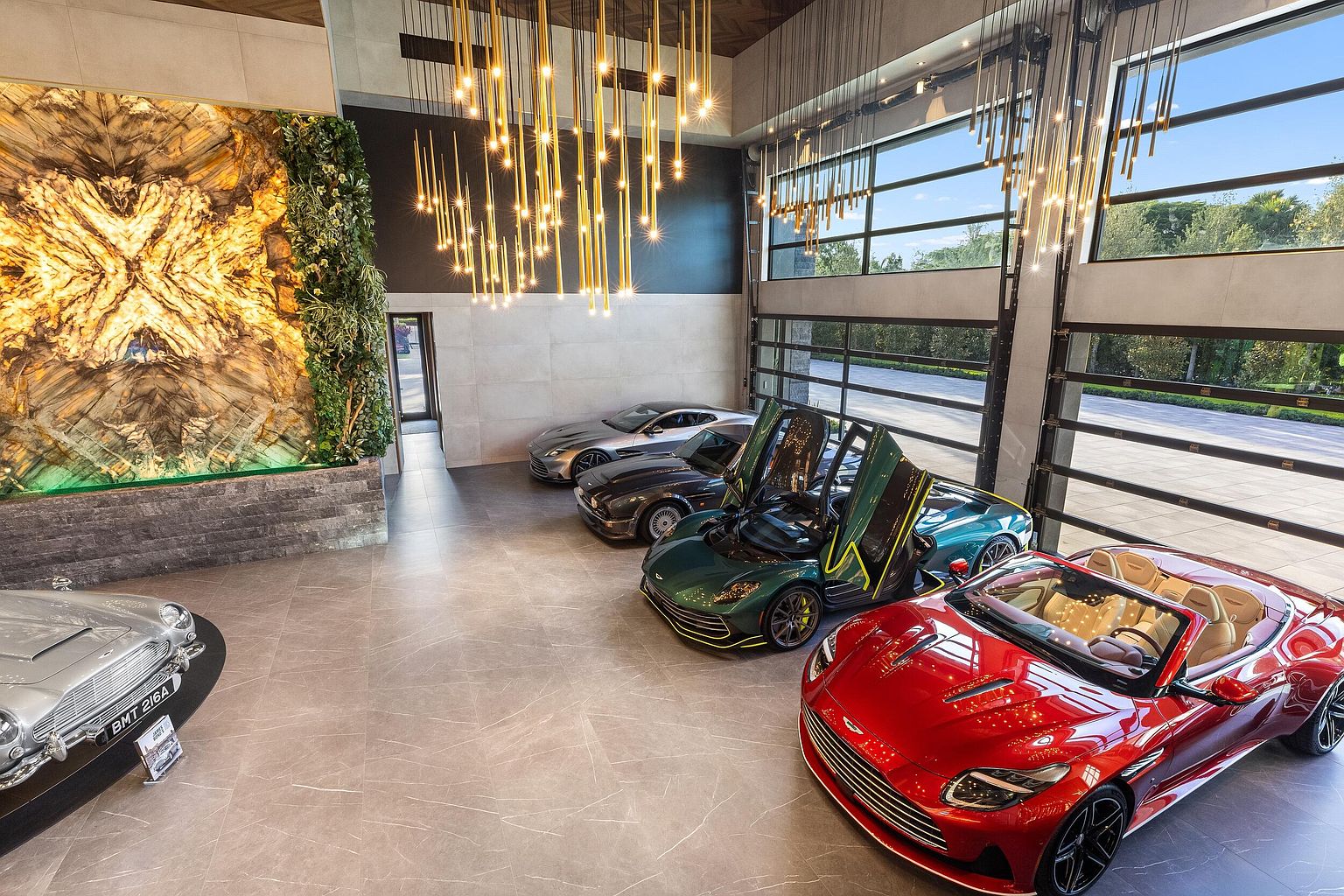 This high-end, climate-controlled garage serves as a luxury showroom for a collection of premium sports cars. The space features large glass garage doors, a dramatic light installation suspended from the ceiling, and a striking illuminated stone feature wall accented by vertical greenery. The perspective is elevated, capturing the polished tile flooring and the curated arrangement of vehicles in a cinematic, gallery-like setting.