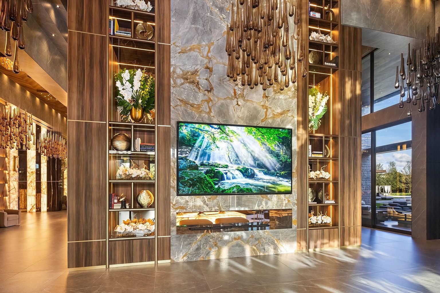 This grand living space features a striking central wall clad in veined stone, housing a large television and a linear fireplace insert. Flanking the central feature are symmetrical, illuminated shelving units crafted from warm-toned wood, filled with decorative accents and greenery. The room is anchored by dramatic, cascading glass pendant light fixtures that create a luxurious, high-end atmosphere.