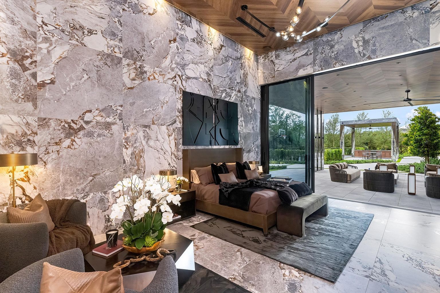 This luxurious primary bedroom features dramatic stone-clad walls and a seamless transition to an expansive outdoor patio through large sliding glass doors. The room is anchored by a modern bed with dark bedding, complemented by a sophisticated seating area and a central light fixture with a contemporary design. The warm wood-paneled ceiling and neutral color palette create an atmosphere of refined elegance and indoor-outdoor connectivity.