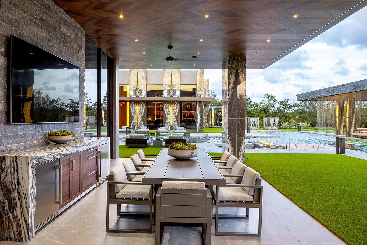 This expansive covered outdoor dining area features a long table with seating for eight, positioned adjacent to a sleek outdoor kitchen with stone countertops and integrated appliances. The space is sheltered by a wood-paneled ceiling with recessed lighting and overlooks a luxurious backyard with a swimming pool, fire feature, and manicured lawn. The design emphasizes a seamless transition between indoor and outdoor living, showcasing high-end finishes and a modern architectural aesthetic.