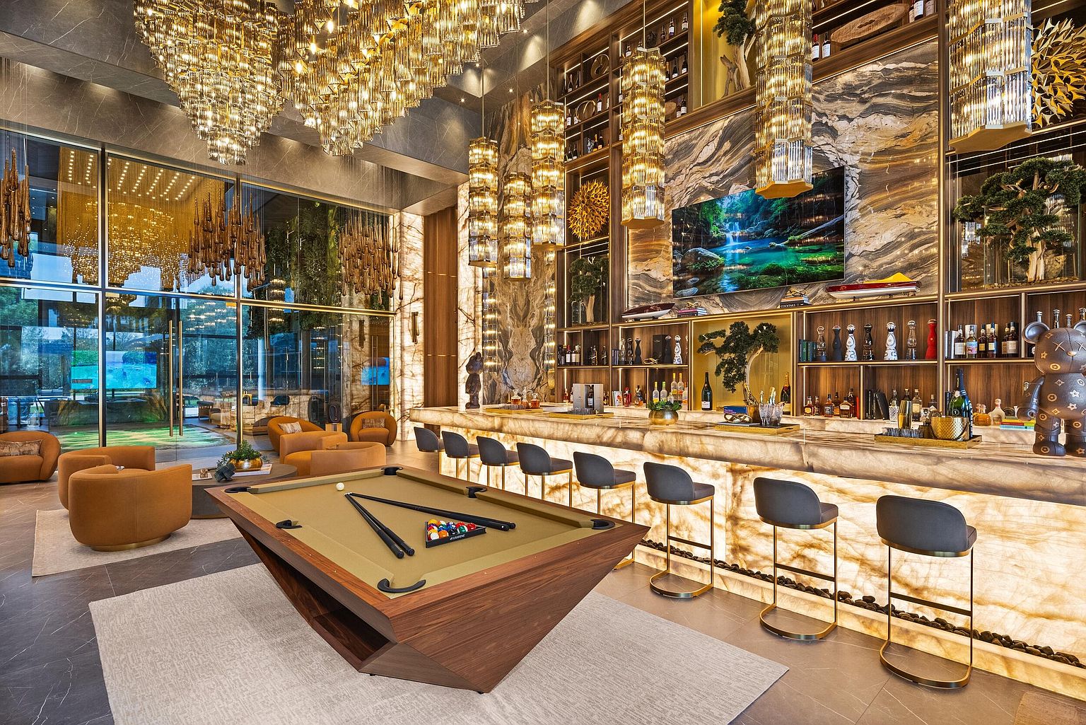 This luxurious lounge area features a high-end bar with a backlit stone counter and a professional-grade pool table as the centerpiece. The space is adorned with elaborate crystal chandeliers, tall windows, and rich wood paneling, creating an opulent atmosphere. The perspective captures the expansive layout, highlighting the sophisticated blend of entertainment and relaxation zones.