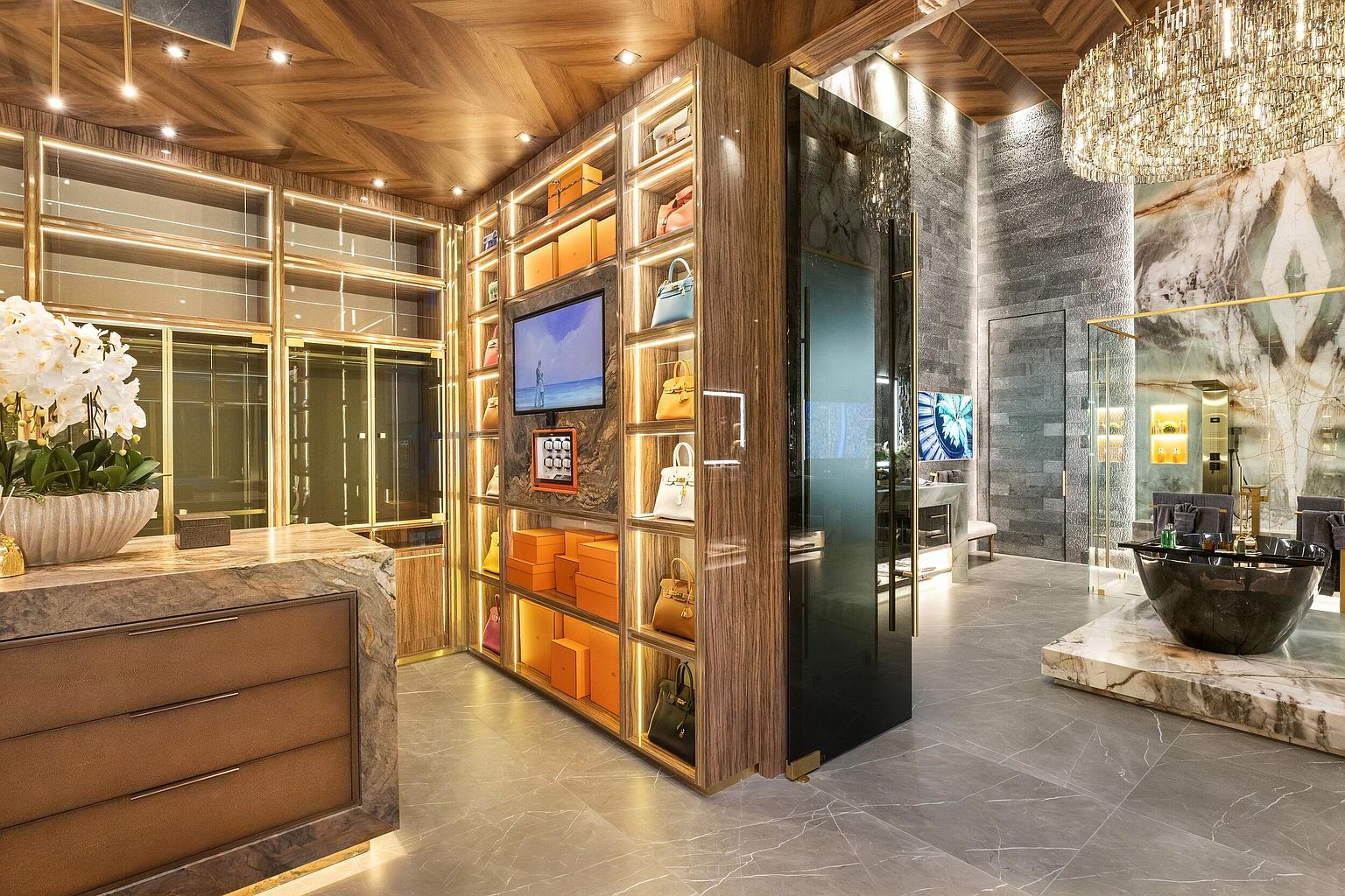 This luxurious walk-in closet features custom wood shelving with integrated LED lighting, showcasing a collection of designer handbags and accessories. The space transitions into a spa-like bathroom area, highlighted by a dark stone soaking tub, a dramatic crystal chandelier, and a large, textured stone feature wall. The combination of rich wood tones, polished stone surfaces, and sophisticated lighting creates an atmosphere of high-end elegance and refined organization.