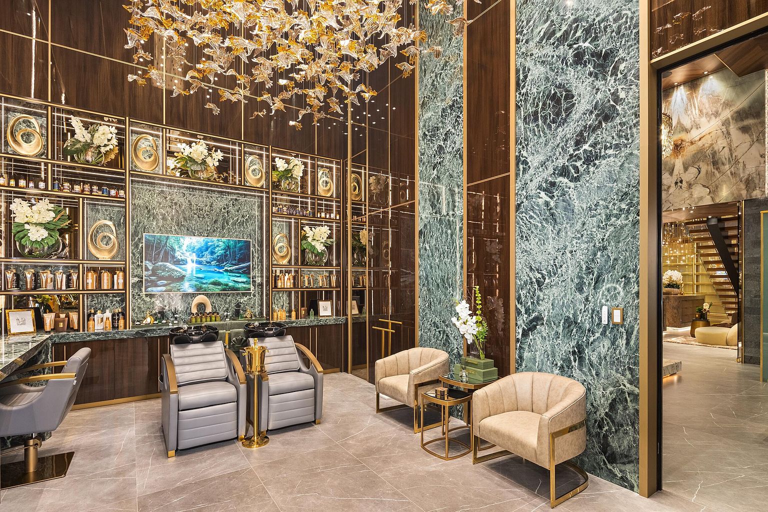 This image showcases a luxurious, high-end salon or spa interior characterized by opulent materials and sophisticated lighting. The space features a prominent green stone wall, dark wood cabinetry with gold-toned metal accents, and a dramatic, sprawling gold-leaf chandelier suspended from the ceiling. Two grey leather reclining chairs are positioned for hair washing, while a pair of cream-colored armchairs with gold frames provide a comfortable seating area, all set against a backdrop of polished stone flooring.